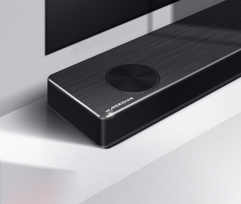 Крупный план левой стороны саундбара LG с лого Meridian в левом нижнем углу. Также виден левый нижний край телевизора.