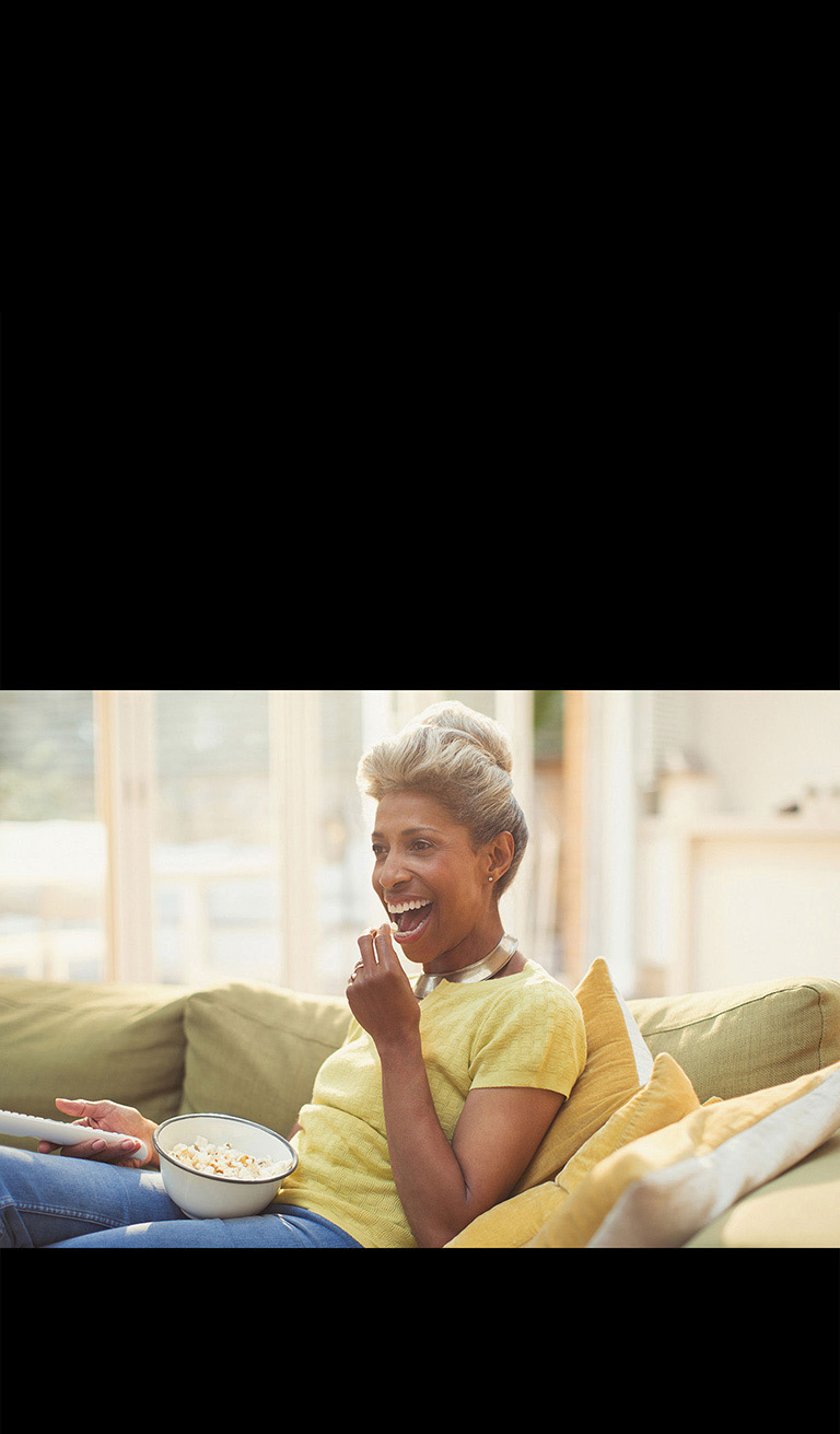 Pult ushlagan bir ayol televizor koʻryapti. U ayni paytda bodroq ham yeyapti.