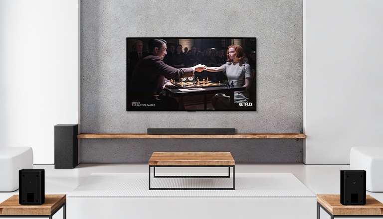Un conjunto de 2 altavoces traseras, un subwoofer y una barra de sonido y un televisor se encuentran en una sala de estar blanca. Una mujer y un hombre juegan al ajedrez en la pantalla del televisor.