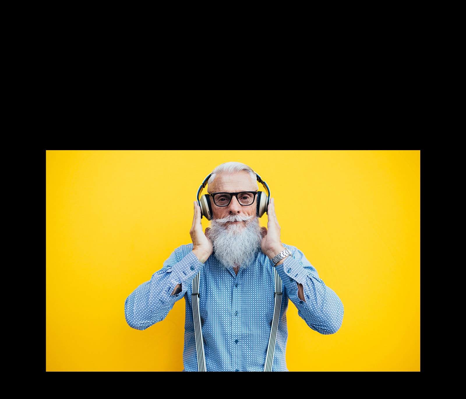 Дедушка в наушника стоит на фоне желтой стены.