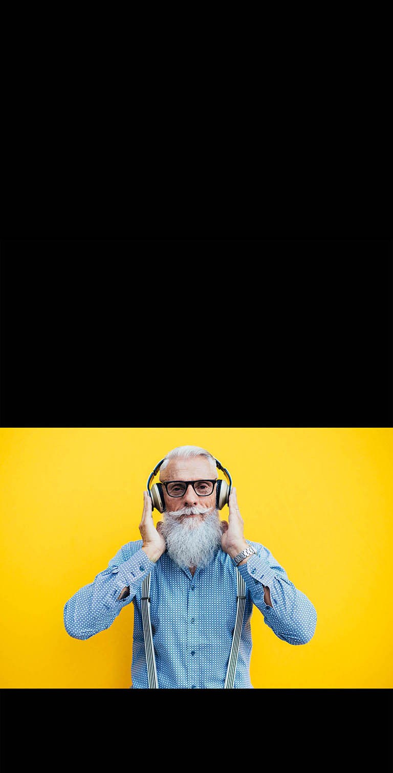 Дедушка в наушника стоит на фоне желтой стены.