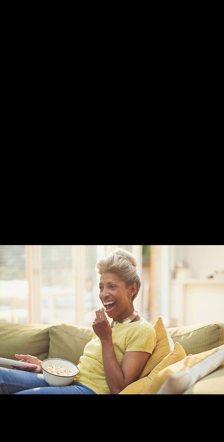 Женщина смотрит телевизор, держа в руках пульт дистанционного управления. Она ест попкорн.