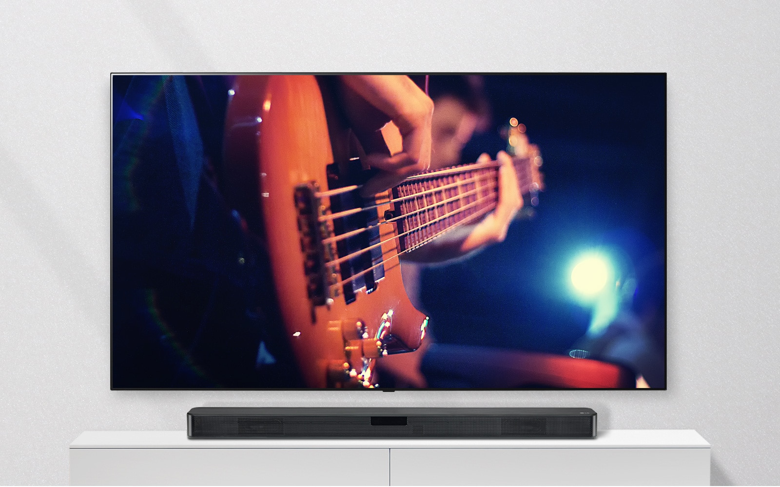TV được gắn vào tường và loa sound bar nằm trên kệ màu trắng. TV hiển thị một người đàn ông đang chơi guitar.