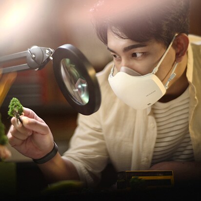 An Asian man is looking at something through a microscope, with a white LG PuriCare™ Wearable Air Purifier on.