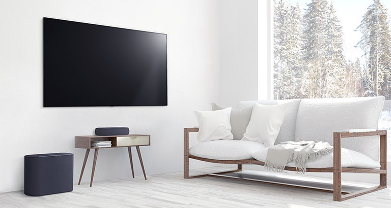 A soundbar is placed on a small tea table,  a subwoofer placed on the left side and TV is placed above a soundbar. The room is overall white. 