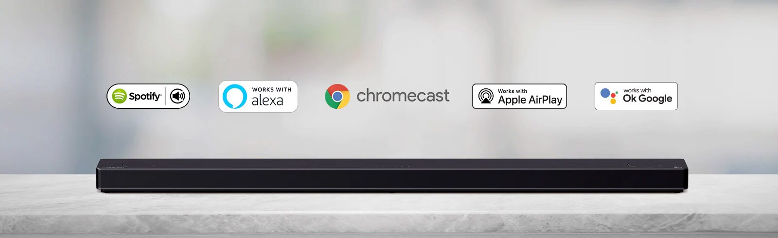 Loa soundbar đặt trên kệ màu xám và có logo nền tảng AI, theo thứ tự spotify, Alexa, Chromecast, Apple Airplay và OK Google từ trái sang phải.