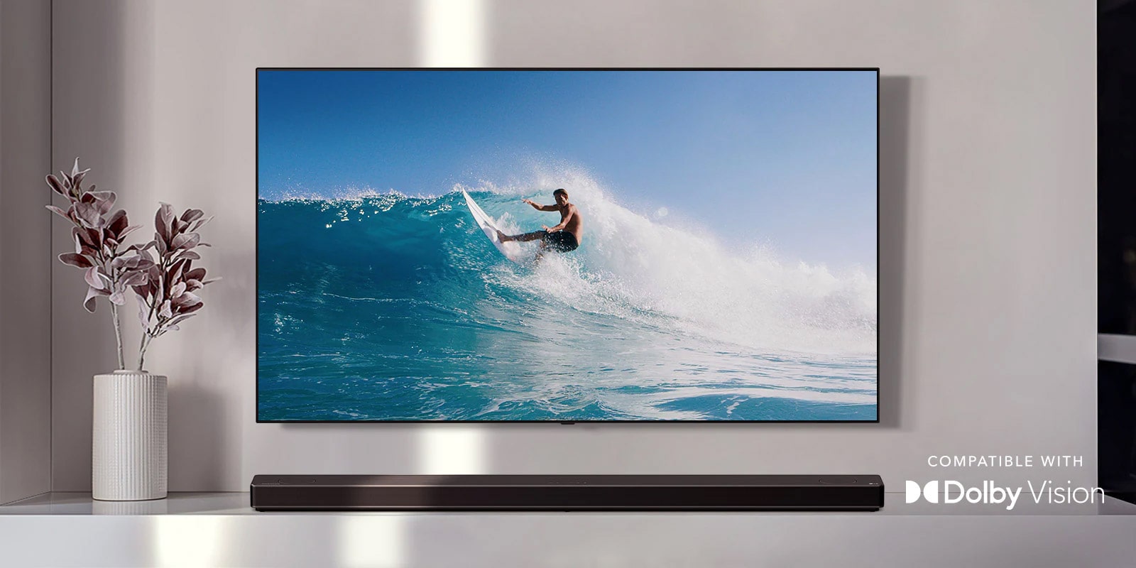 TV treo trên tường. Tv thể hiện một người đàn ông đang lướt trên con sóng lớn. LG Soundbar nằm ngay bên dưới TV trên kệ trắng. Một chiếc bình với một bông hoa ngay bên cạnh loa soundbar. (phát video)