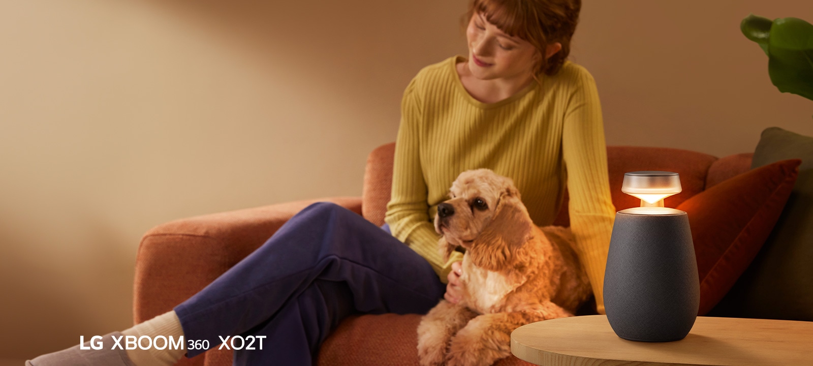 Một người phụ nữ ngồi trên ghế sofa với chú chó của cô và nghe nhạc bằng LG XBOOM 360 XO2T.