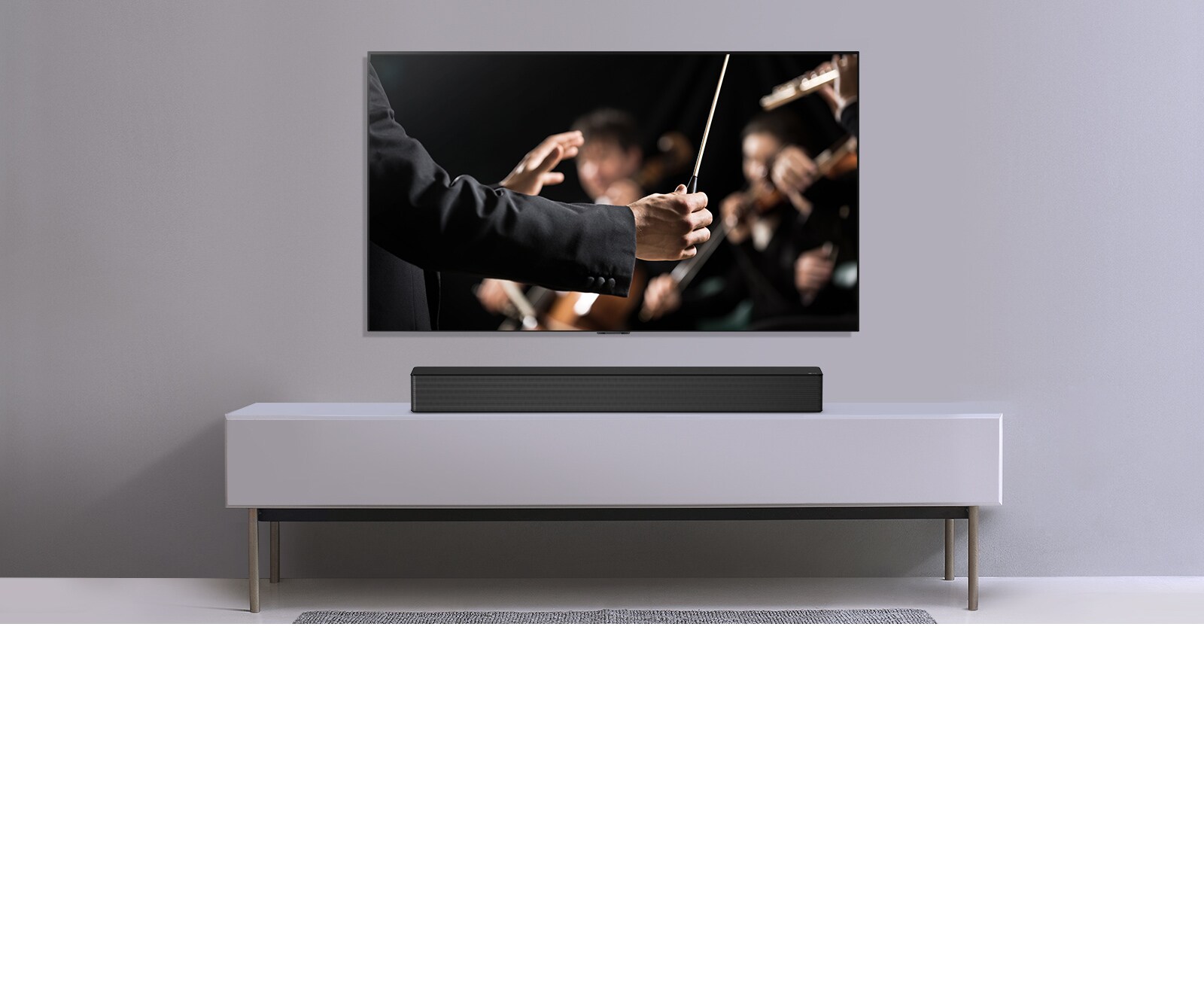 TV được hiển thị trên một bức tường màu xám và Loa thanh LG đặt bên dưới trên kệ màu xám. TV hiển thị một nhạc trưởng đang điều khiển dàn nhạc.