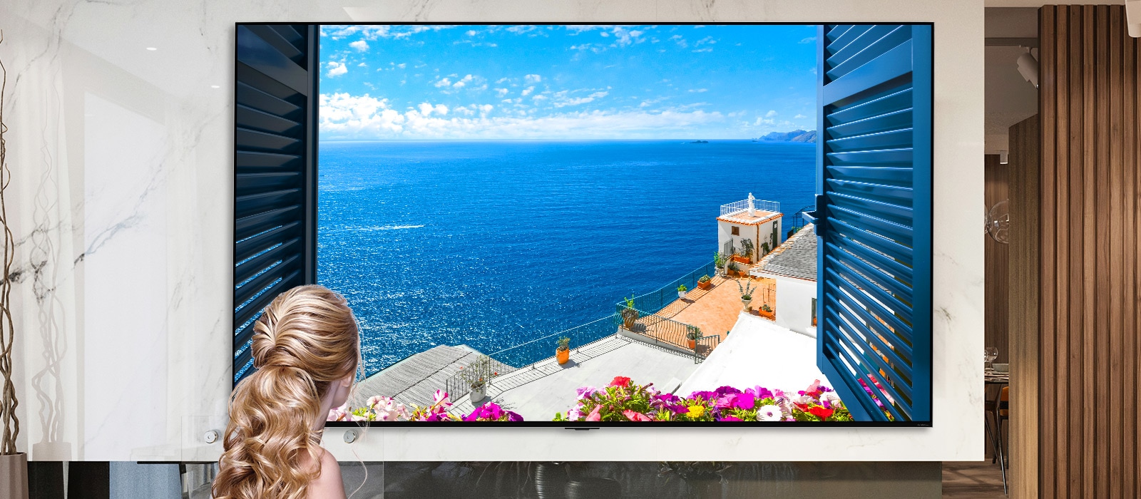 Rear view of a woman looking out of a window at a blue sea and white buildings. As the page scrolls down the image zooms out to show more of the room and reveal the window to be an LG QNED MiniLED TV.