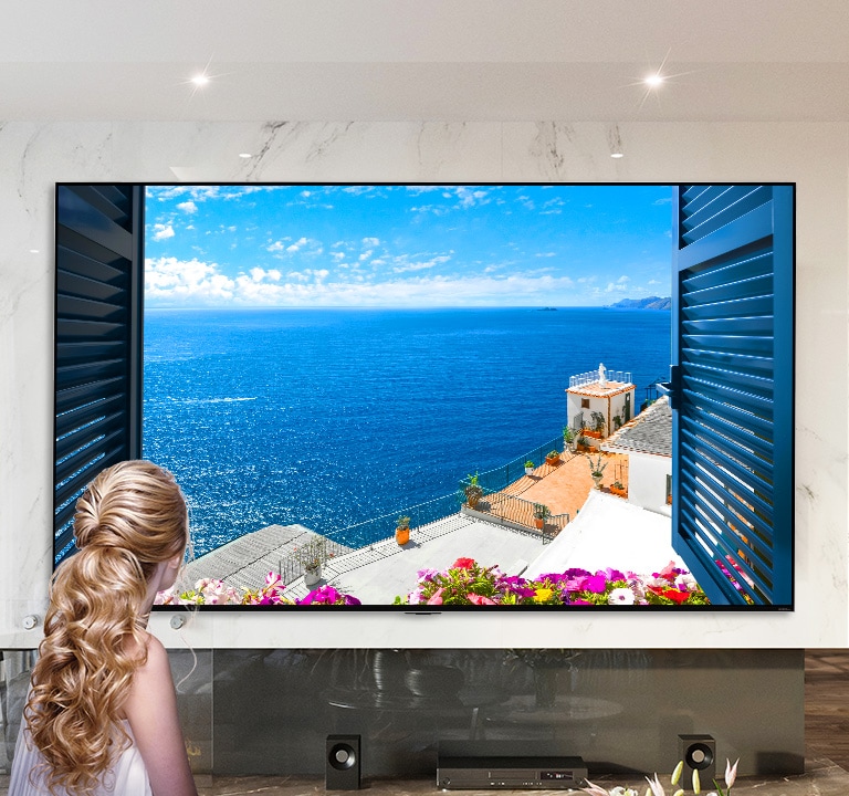 Rear view of a woman looking out of a window at a blue sea and white buildings. As the page scrolls down the image zooms out to show more of the room and reveal the window to be an LG QNED MiniLED TV.