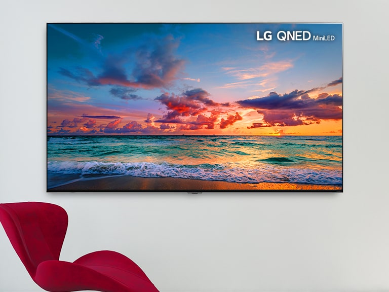 A large-screen wall mounted TV with a red chair in front. The screen shows waves gently breaking on a beach. Scrolling from left to right shows the difference in color on a conventional LCD TV versus LG QNED MiniLED.