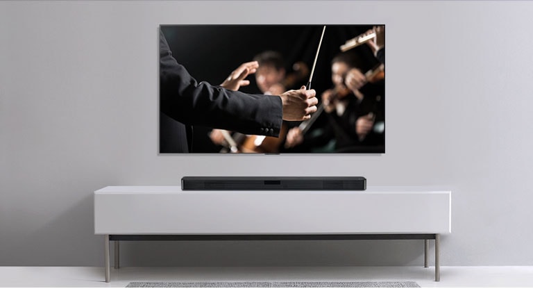 É mostrada uma TV numa parede cinza, com a LG Soundbar por baixo numa prateleira cinza. A TV mostra um maestro conduzindo uma orquestra.
