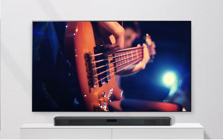 A TV está montada na parede e o sound bar está numa prateleira branca. TV a mostrar um homem a tocar guitarra.