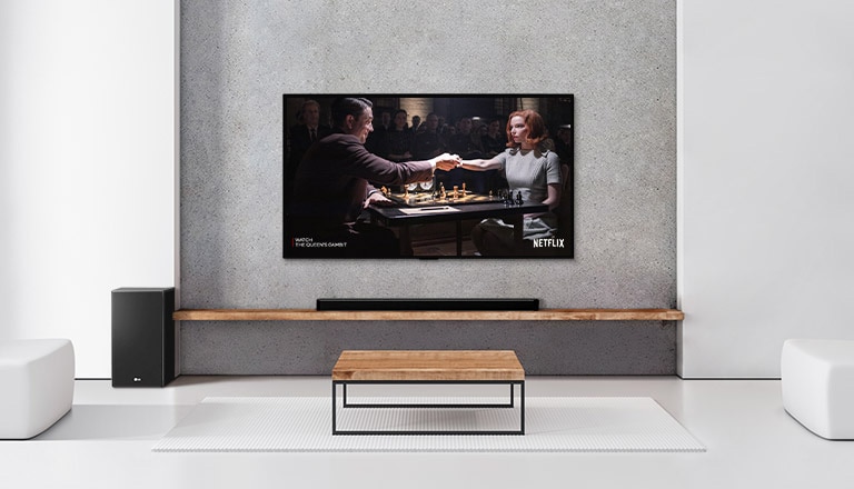 Um subwoofer, uma barra de som e uma TV estão em uma sala de estar branca. Uma mulher e um homem jogam xadrez na tela da televisão.