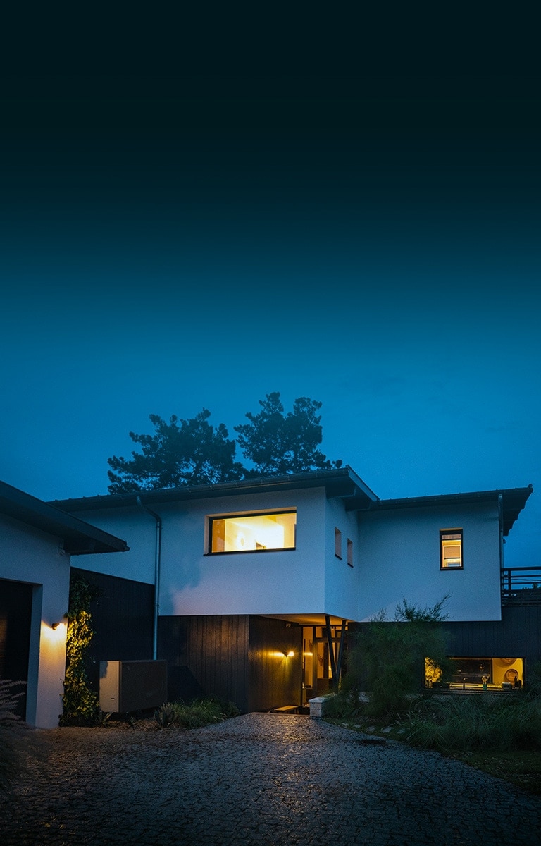 Gesamtes Äußeres eines Hauses mit warmem Licht an einem kalten Winterabend. Vor dem Haus sehen wir die neue schwarze Luft-Wasser-Wärmepumpe THERMA V von LG. 