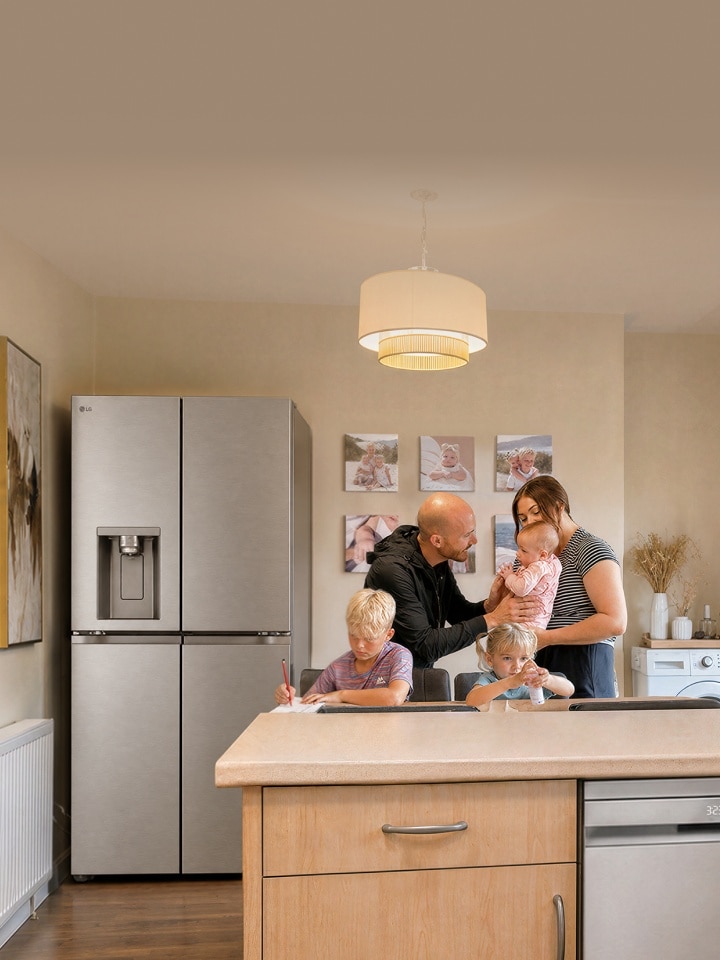 Une famille réunie dans une cuisine, près d'un réfrigérateur LG argenté : un garçon écrit à la table à manger, une petite fille est assise à côté de lui avec une petite bouteille, et les parents se tiennent ensemble en portant un bébé.
