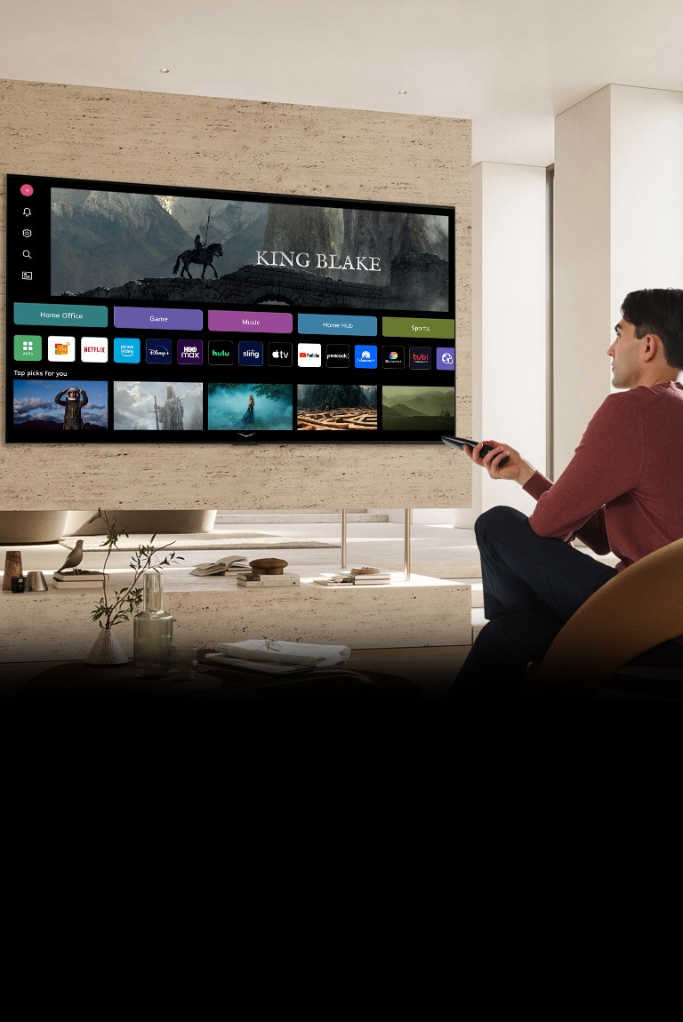 Un hombre sostiene un control remoto en su mano derecha, mirando un televisor grande frente a él. La pantalla del televisor muestra la pantalla "Nuevo hogar".