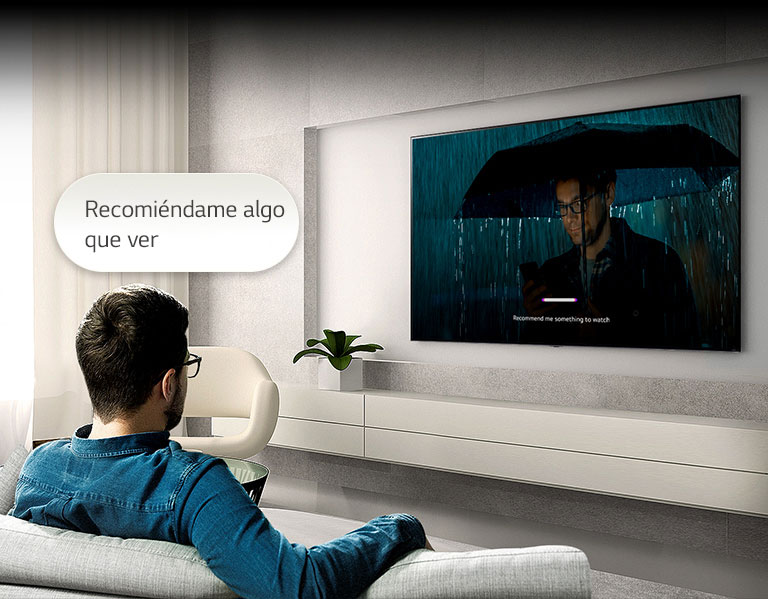 Un hombre sentado en un sofá frente a un televisor. Una burbuja de diálogo que dice "Recomiéndame algo que ver" flota sobre su cabeza. Dentro de la pantalla, se reproduce un video de un hombre con un paraguas con una interfaz de usuario que se reconoce por voz.