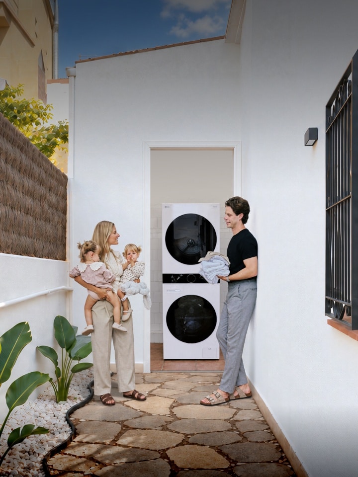 A family with toddler twins standing in a white-walled outdoor hallway next to a stacked LG washing machine and tumble dryer unit.