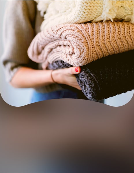 Person holding a stack of folded knitted clothes in neutral colors, including cream, beige, and dark gray sweaters