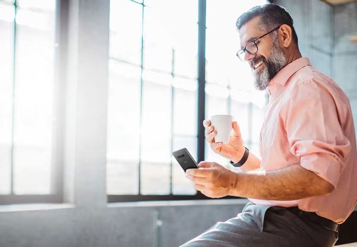 A man holding onto a mug whilst smiling at his LG Smartphone.