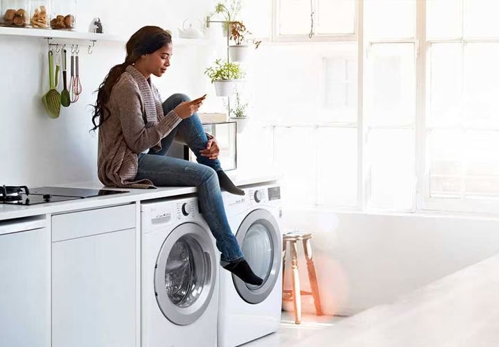A woman on her phone whilst sitting on top of her LG washing machine and dryer.