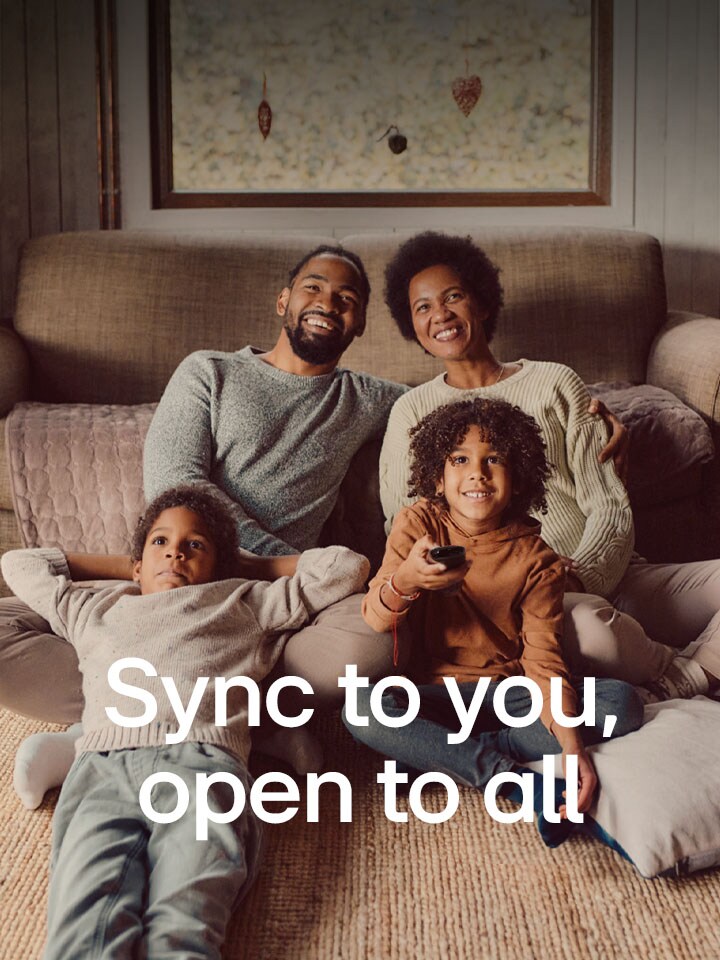 An image of a family of four sitting on the floor in front of cozy brown sofa looking straight ahead and laughing while watching TV. The words "Syncs to you, open to all" overlay the image in a white font. 