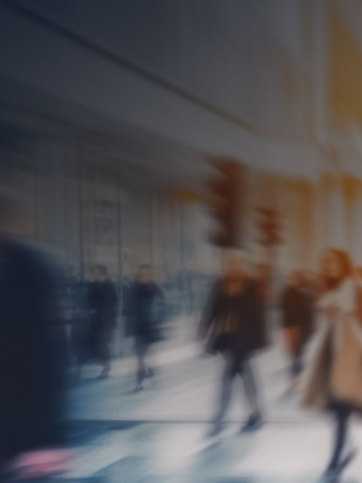 Blurred figures of people walking on a busy city street in daylight, suggesting motion and urban activity.