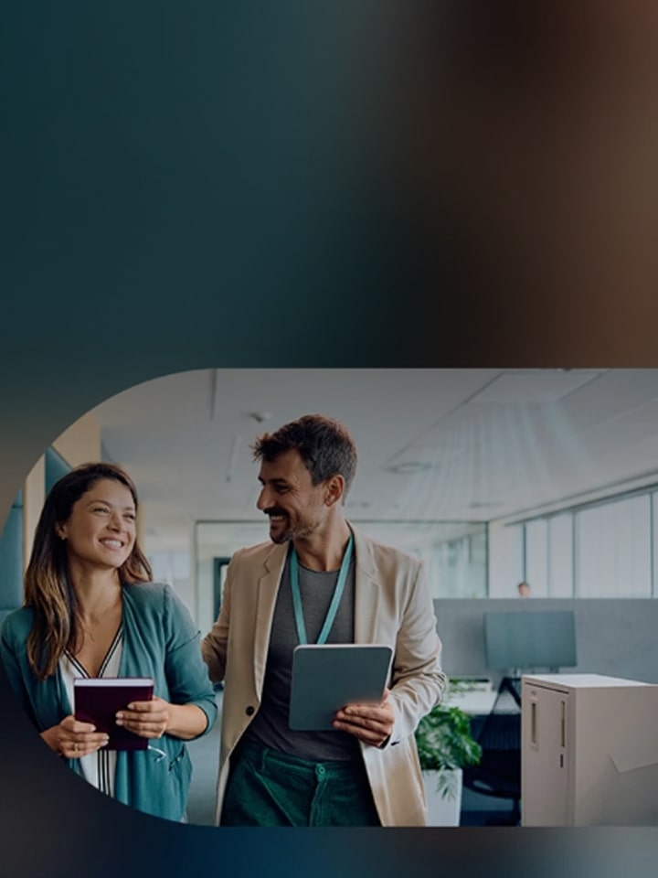 Two smiling coworkers walking together in an office, holding a notebook and a tablet while having a friendly conversation.