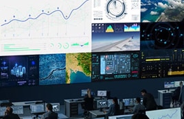 Kontrollraum mit mehreren großen Bildschirmen, auf denen Datendiagramme, Karten und Überwachungs-Dashboards angezeigt werden, während die Mitarbeiter an Computerarbeitsplätzen arbeiten.