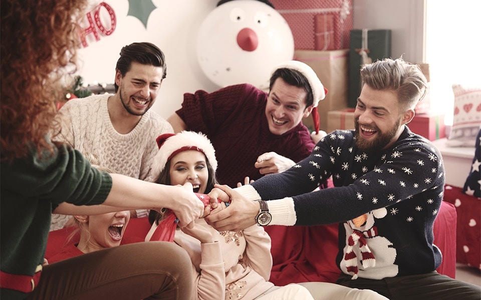 A group of six friends laugh and smile while playing a Christmas game in a holiday-themed room