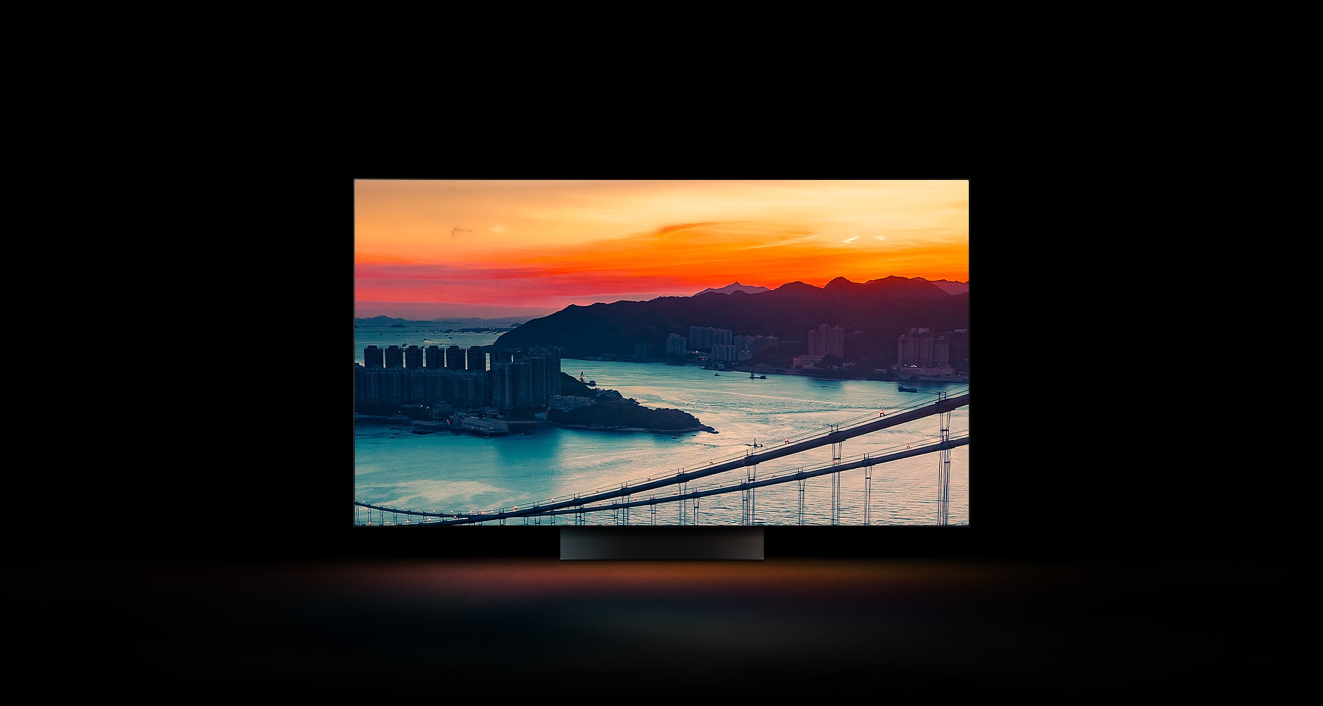 An ariel view of a coastal city with traffic driving over a bridge as the sun sets appears across the full screen. It is cropped and shown on an LG OLED television screen with the original colours unchanged.