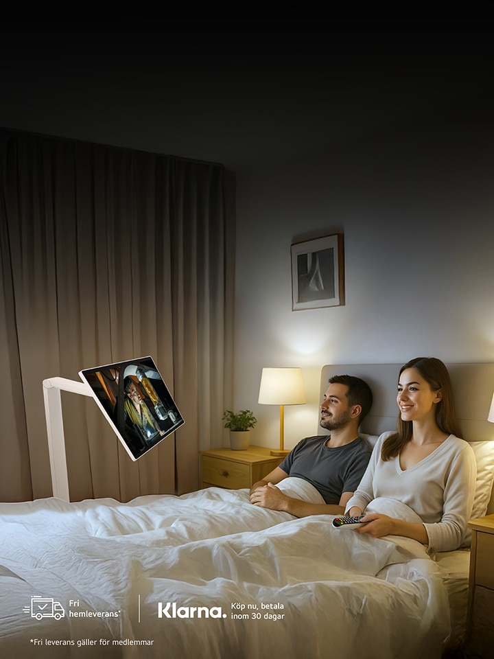Ett par ligger i sängen och tittar på en LG Smart Monitor som är monterad på en Swing-arm framför dem. Rummet är mjukt upplyst med en sänglampa, och skärmen visar färgglatt innehåll. Båda ser avslappnade ut. Längst ner i bilden syns logotyperna för LG och Swing Smart Monitor.