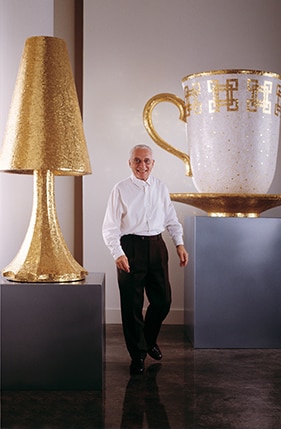 Alessandro Mendini im Bild zusammen mit seiner künstlerischen Skulpturen in Form einer Tasse und einer Lampe.