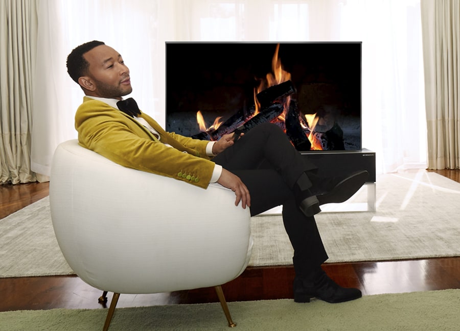 John Legend sitzt in einem weißen Stuhl vor einem aufrollbaren OLED-Fernseher von LG SIGNATURE.