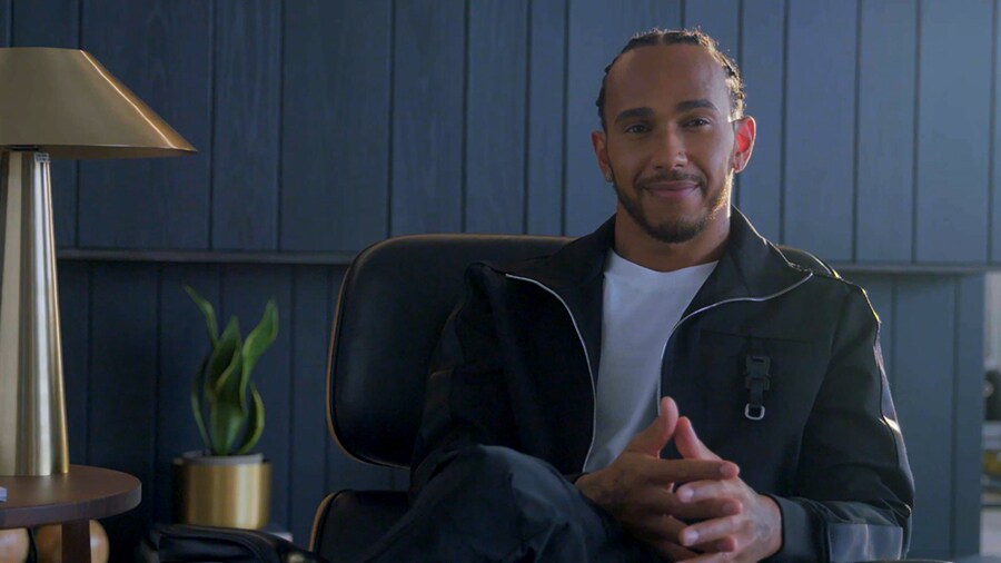Lewis Hamilton is sitting and resting his arms on a black armchair with his legs crossed.