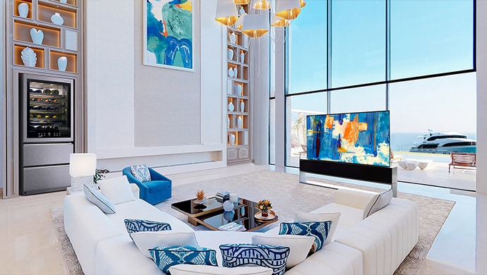 LG SIGNATURE Rollable OLED TV R and Wine Cellar in a white-and-blue-themed room in front of large windows with a view of the ocean.
