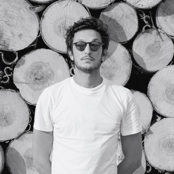 Foto in bianco e nero di Delfino Sisto Legnani in piedi davanti a una catasta di legname.