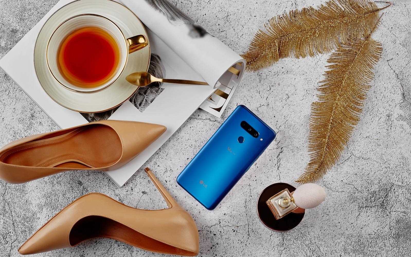 Stylowy smartfon LG V40 w otoczeniu złotych piór, eleganckich szpilek, filiżanki z herbatą i perfum